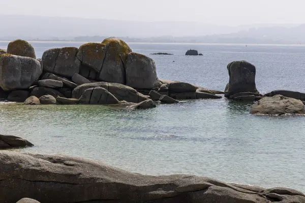 Arka planda büyük bir su kütlesi olan kayalık bir kıyı şeridi. Kıyı boyunca dağılan kayalar engebeli ve doğal bir manzara yaratıyor. Su sakin görünüyor..