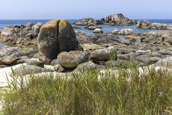 Arka planda küçük bir su kütlesi olan kayalık bir kıyı şeridi. Kayalar bölgenin dört bir yanına dağılmış, bazıları daha büyük, bazıları daha küçük. Gökyüzü açık ve mavi