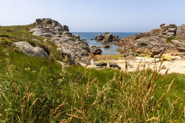 Önünde küçük bir su kütlesi olan kayalık bir sahil. Plaj çoğunlukla çimlerle kaplıdır ve bazı kayalar etrafa saçılmıştır. Gökyüzü açık ve mavidir ve güneş ışıl ışıl parlar.