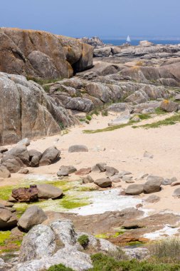 Uzakta küçük bir yelkenli olan kayalık bir sahil. Kayalar sahile saçılmış ve gökyüzü açık ve mavi. Huzurlu ve huzurlu bir sahne.