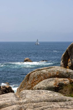 Bir yelkenli okyanusta kayaların yanında seyrediyor. Gökyüzü açık ve su sakin.