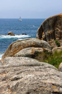 Bir yelkenli okyanusta kayaların yanında seyrediyor. Gökyüzü açık ve su sakin.