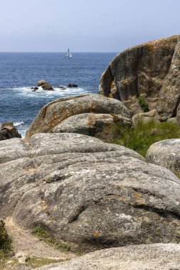 Bir yelkenli okyanusta kayaların yanında seyrediyor. Gökyüzü açık ve su sakin.