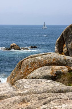 Bir yelkenli okyanusta kayaların yanında seyrediyor. Gökyüzü açık ve su sakin.