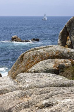 Bir yelkenli okyanusta kayaların yanında seyrediyor. Gökyüzü açık ve su sakin.