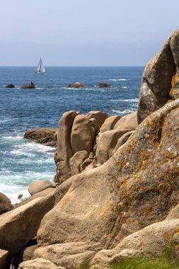Bir yelkenli okyanusta kayaların yanında seyrediyor. Gökyüzü açık ve su sakin.