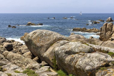 Bir yelkenli okyanusta kayaların yanında seyrediyor. Gökyüzü açık ve su sakin.