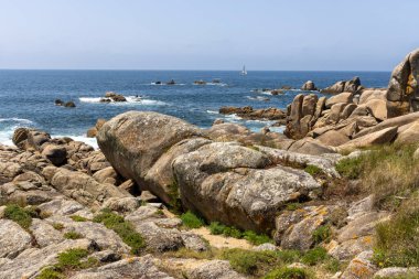 Bir yelkenli okyanusta kayaların yanında seyrediyor. Gökyüzü açık ve su sakin.