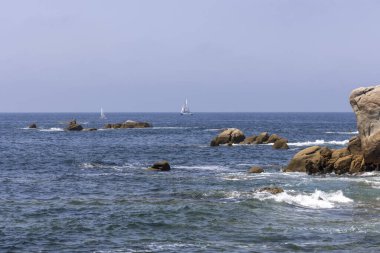 Bir yelkenli okyanusta kayaların yanında seyrediyor. Gökyüzü açık ve su sakin.
