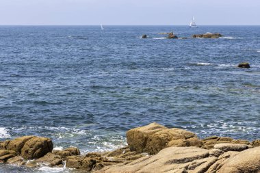 Bir yelkenli okyanusta kayaların yanında seyrediyor. Gökyüzü açık ve su sakin.