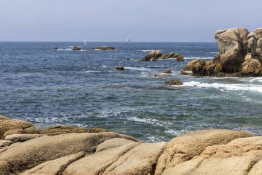Bir yelkenli okyanusta kayaların yanında seyrediyor. Gökyüzü açık ve su sakin.