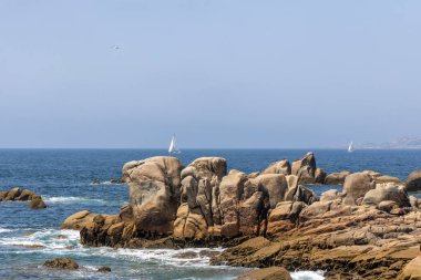 Bir yelkenli okyanusta kayaların yanında seyrediyor. Gökyüzü açık ve su sakin.