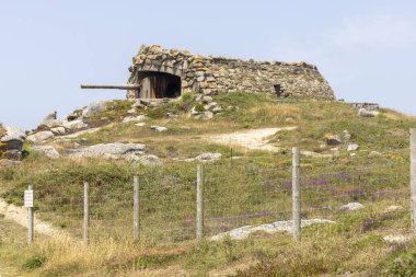 Tepesinde küçük bir bina olan kayalık bir tepe. Bina taştan yapılmış ve tepesinde bir top var. Tepe kayalarla çevrili ve ön planda çok fazla ot var.