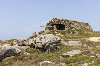 Tepesinde küçük bir bina olan kayalık bir tepe. Bina taştan yapılmış ve tepesinde bir top var. Tepe kayalarla çevrili ve ön planda çok fazla ot var.