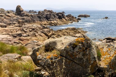 Önünde büyük bir kaya olan kayalık bir kıyı şeridi. Kayalar kıyı şeridi boyunca dağılmış ve su sakin. Sahnenin huzurlu ve huzurlu bir havası var.