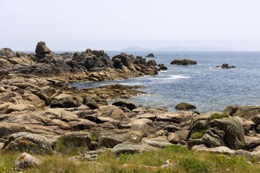 Önünde büyük bir kaya olan kayalık bir kıyı şeridi. Kayalar kıyı şeridi boyunca dağılmış ve su sakin. Sahnenin huzurlu ve huzurlu bir havası var.