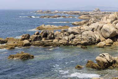 Arka planda sakin bir okyanus olan kayalık bir kıyı şeridi. Kayalar sahile dağılmış, engebeli ve doğal bir manzara yaratıyorlar. Huzur ve sükunet kavramı