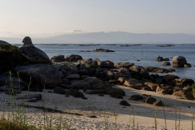 Önünde büyük bir kaya olan kayalık bir sahil ve arka planda midye çiftlikleri plaj boş ve gökyüzü bulutlu.