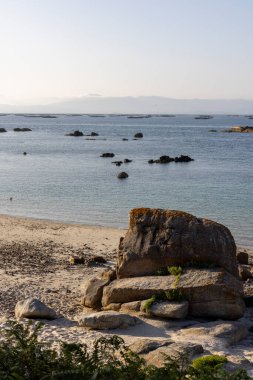 Önünde büyük bir kaya olan kayalık bir sahil. Okyanus sakin ve gökyüzü berrak