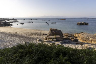 Önünde büyük bir kaya olan kayalık bir sahil. Okyanus sakin ve gökyüzü berrak