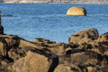 Büyük bir kaya bir su kütlesinin ortasında oturur. Su sakin ve gökyüzü berrak. Kaya kayalarla çevrili ve manzara huzur dolu.
