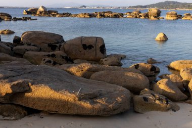 Büyük bir kaya bir su kütlesinin ortasında oturur. Su sakin ve gökyüzü berrak. Kaya kayalarla çevrili ve manzara huzur dolu.