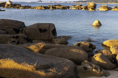 Büyük bir kaya bir su kütlesinin ortasında oturur. Su sakin ve gökyüzü berrak. Kaya kayalarla çevrili ve manzara huzur dolu.