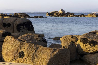 Büyük bir kaya bir su kütlesinin ortasında oturur. Su sakin ve gökyüzü berrak. Kaya kayalarla çevrili ve manzara huzur dolu.