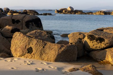 Büyük bir kaya bir su kütlesinin ortasında oturur. Su sakin ve gökyüzü berrak. Kaya kayalarla çevrili ve manzara huzur dolu.