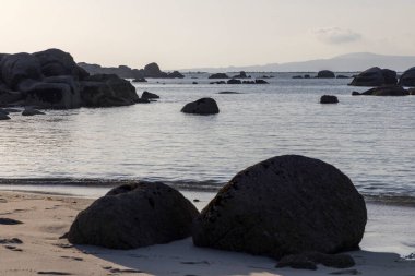 Önünde büyük bir kaya olan kayalık bir kıyı şeridi. Su sakin ve gökyüzü açık mavi.
