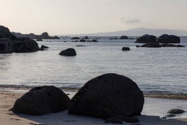 Önünde büyük bir kaya olan kayalık bir kıyı şeridi. Su sakin ve gökyüzü açık mavi.