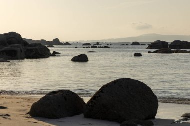 Önünde büyük bir kaya olan kayalık bir kıyı şeridi. Su sakin ve gökyüzü açık mavi.