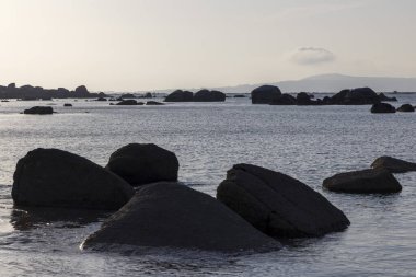 Önünde büyük bir kaya olan kayalık bir kıyı şeridi. Su sakin ve gökyüzü açık mavi.