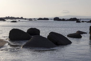 Önünde büyük bir kaya olan kayalık bir kıyı şeridi. Su sakin ve gökyüzü açık mavi.
