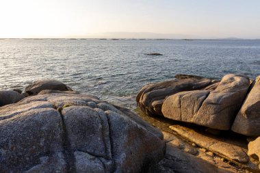 Önünde büyük bir kaya olan kayalık bir kıyı şeridi. Su sakin ve gökyüzü açık mavi.