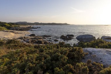 Arka planda su bulunan kayalık bir sahil. Gökyüzü mavi ve güneş batıyor.