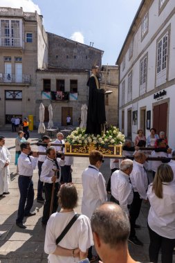 Allariz, İspanya, 13 Temmuz 2025: Büyük bir heykel taşıyan bir grup insan. İnsanlar siyah ve beyaz giyinmiş ve heykel çiçeklerle çevrili..