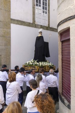 Allariz, İspanya, 13 Temmuz 2025: Büyük bir heykel taşıyan bir grup insan. İnsanlar siyah ve beyaz giyinmiş ve heykel çiçeklerle çevrili..