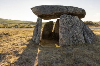 Ortasında küçük bir delik olan büyük bir kaya yapısı. Güneş kayaların üzerinde parlıyor, sıcak ve davetkar bir atmosfer yaratıyor.