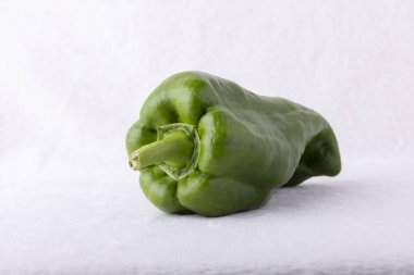 Beyaz arka planda yeşil bir biber duruyor. Biber büyük ve bir sapı dışarı yapışmış. Tazelik ve doğal güzellik kavramı