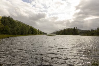 Arka planda bulutlu bir gökyüzü olan sakin bir göl. Su durgun ve gökyüzü kapalı.