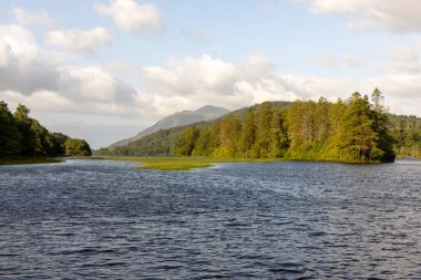 Arka planda dağ olan sakin bir göl. Su durgun ve berrak. Gökyüzü bulutlu, ama güneş bulutların arasından parlıyor. Huzurlu ve huzurlu bir sahne.