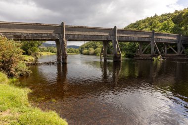 Arka planda bulutlu bir gökyüzü olan bir nehir boyunca uzanır. Su sakin ve köprü eski.