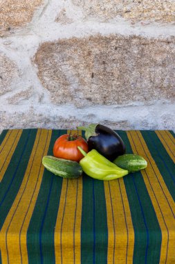 Yeşil ve sarı çizgili bir masa örtüsünde domates, salatalık ve dolma biber gibi çeşitli sebzeler sıralanır.