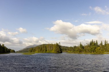 Arka planda birkaç ağacın olduğu sakin bir göl. Gökyüzü çoğunlukla açık ve birkaç bulut dağılmış.