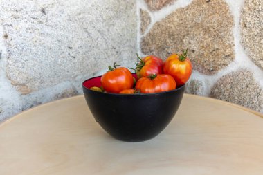 Bir kase domates masada oturur. Kâse siyah ve domatesler kırmızı. Kase domateslerle dolu ve hepsi farklı boyutlarda.