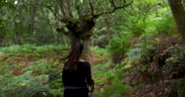 Kadının ve iki köpeğinin eğrelti otlarıyla yemyeşil bir ormanda yürürken çekilmiş bir fotoğrafı. Kadın siyah gömlek ve pantolon giyiyor..