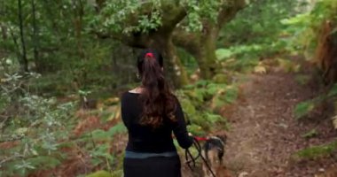 Kadının ve iki köpeğinin eğrelti otlarıyla yemyeşil bir ormanda yürürken çekilmiş bir fotoğrafı. Kadın siyah gömlek ve pantolon giyiyor..