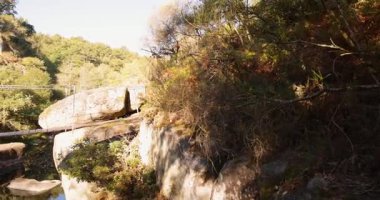 A bridge over a river with a forest in the background. The bridge is wooden and has a metal railing. The water is calm and the sky is clear. Pan left.