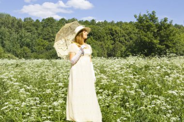 Beyaz tarihi elbiseli, beyaz şemsiyeli genç bir kadın. Çiçekli çayırda, yaban havucu çiçekleri ve ormanla birlikte..